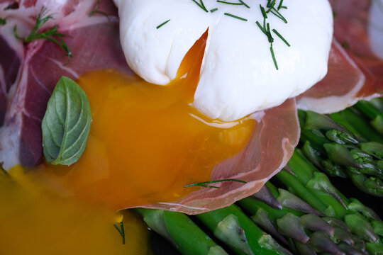 A dish is prepared with poached egg, green asparagus, and thinly sliced ham. The food is arranged on a black plate. This scene is set in a kitchen. Healthy eating concepts.