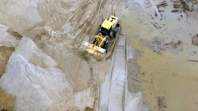 Aerial view shows bulldozer moving through sand piles. Machine scrapes and pushes loose sand sideways. Large dust trail follows the equipment's path. Active mining or land reshaping work