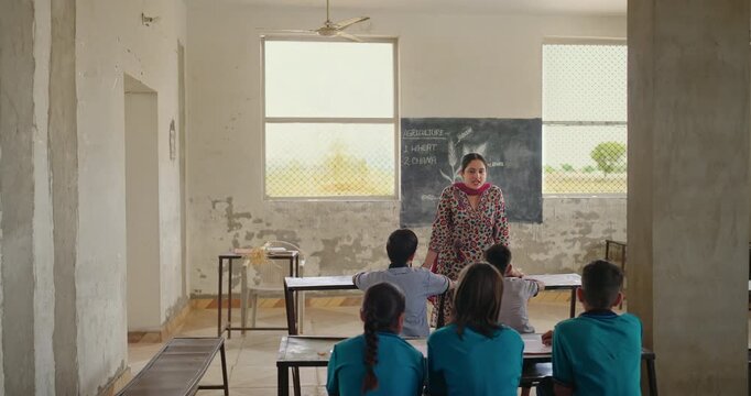 Indian young adult villager female talk speak to child group indoor class room rural area village place teen boy girl sitting on bench wear uniform look tutor do study learn raise hand day time 