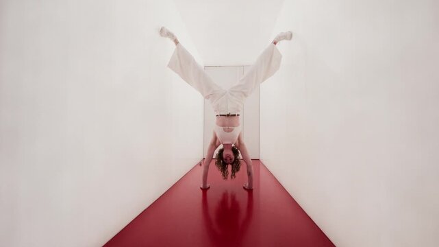Full portrait of young Caucasian woman in white outfit performing handstand with split legs in minimalist corridor with red floor. Suitable for fitness, mental strength, and balance concepts