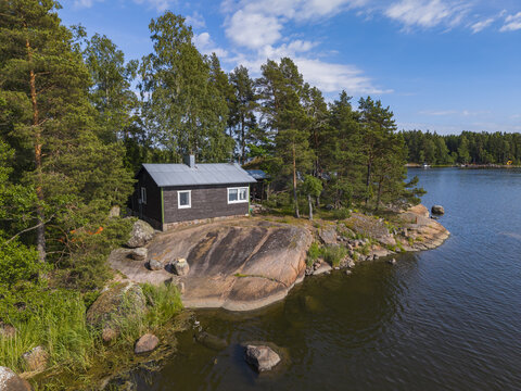 Aerial view of a small wooden cabin nestled on a rocky shoreline surrounded by pine trees and calm lake waters in Hamina, Kymenlaakso, Finland.