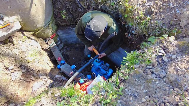 A person works to fix a water pump located in a hole in a yard. Tools are scattered nearby. The ground is disturbed with soil and rocks visible around the area. The worker is focused on connecting pip