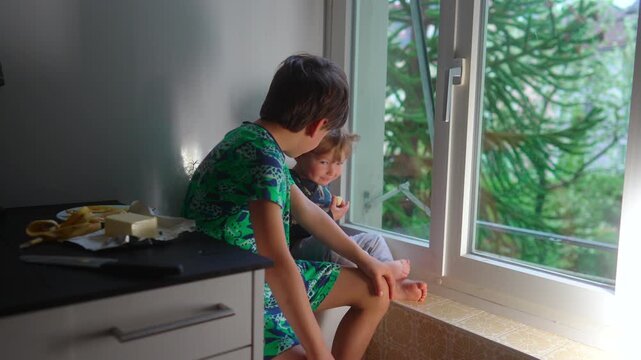 Toddler enjoying a banana snack beside older brother during a calm morning at home highlighting sibling companionship emotional security and everyday family connection