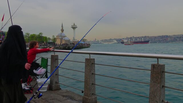 A woman wearing a black niqab holds a fishing rod while sitting on the stone embankment of Uskudar overlooking the Bosphorus sea and mosque in Istanbul