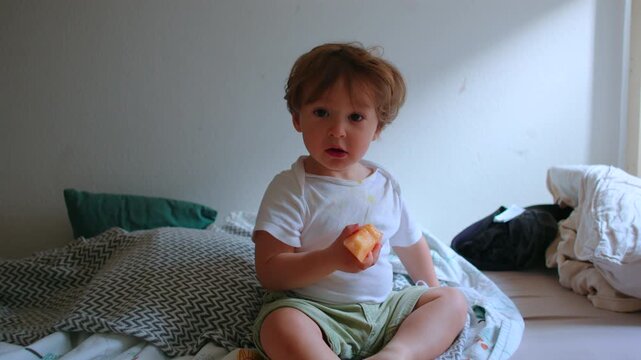 Toddler sitting on bed eating melon slice during relaxed snack time, looking toward camera with curious expression, healthy childhood nutrition and everyday family life at home