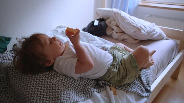 Toddler relaxing on bed while eating fresh melon slice, comfortable home moment highlighting healthy childhood nutrition, independence and everyday family life indoors