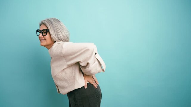 Senior woman with grey hair experiencing sudden lower back pain. She is wincing, holding her back, and expressing discomfort and suffering