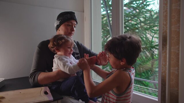 Mother caressing older son while he playfully claps toddler&rsquo;s hands together, authentic family interaction expressing affection, sibling bonding, parental love and emotional connection at home