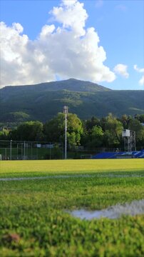 Football stadium timelapse with moving clouds above green field and distant mountains on a sunny day, dynamic sky changing over sports arena, outdoor landscape with dramatic weather, scenic stadium