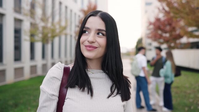 Pretty female student with a backpack smiling and looking at the camera. She stands in the university campus yard with friends in the background