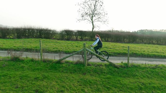 Female cyclist pedaling along countryside path near green farmland and hedgerows, slow motion. Healthy outdoor lifestyle workout improving stamina endurance, recreational cycling activity. Rural