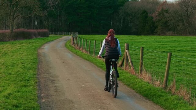 Middle aged woman cycling countryside road between green meadows and forest edge enjoying outdoor fitness. Active female cyclist riding bicycle along rural lane during calm daylight promoting healthy