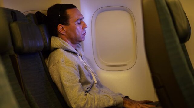 Weary passenger dozing in economy class seat, head nodding forward under soft bluish glow from in-flight screen. Suddenly waking up, man immediately looks at screen and starts tapping it. Side view