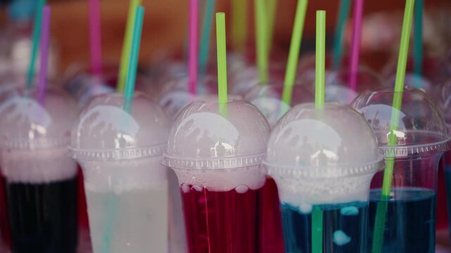 Colorful fizzy drinks bubbling in plastic cups with with dry ice straws at outdoor market stall slow tracking pan across rows.