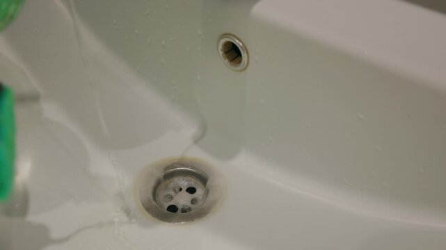 Cleaning with acid, gloved hand scrubbing bathroom sink basin with sponge to remove stains and reveal clean porcelain drain area.