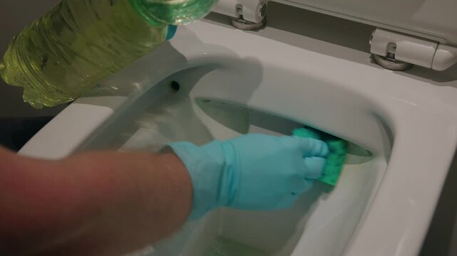 Cleaning with acid, Handheld close up of gloved hand scrubbing toilet bowl rim inside bathroom with liquid cleaner.