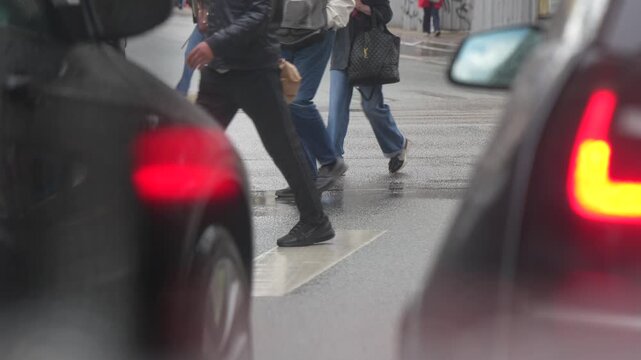 Foot traffic navigating a damp pedestrian street crossing with glowing red vehicle brake lights in the foreground, representing urban lifestyle