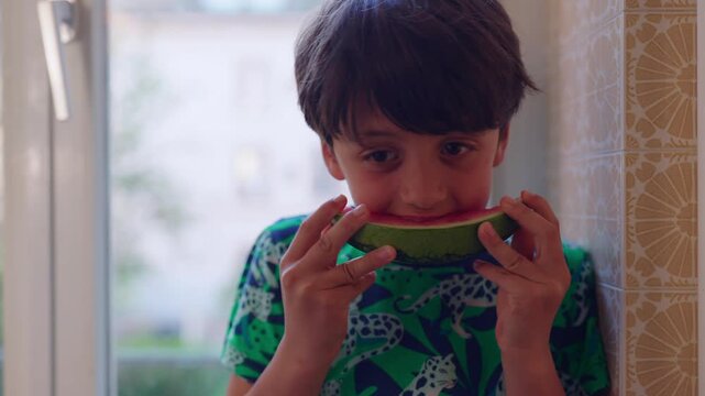 Young boy eating watermelon slice by window at home, healthy childhood nutrition moment highlighting wellbeing, freshness, enjoyment and natural eating habits