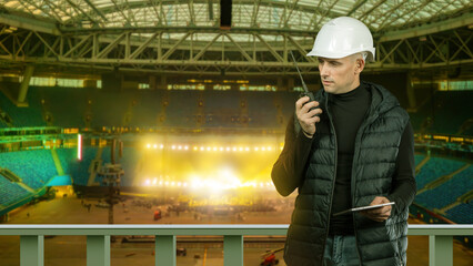 Stadium event production. Site manager coordinates stage setup and lighting installation inside large arena using radio and tablet © Grispb