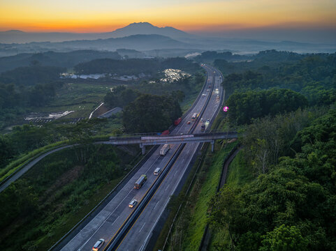 Aerial view of Jalan Tol Bawen - Salatiga highway winding through lush green hills and rice fields at sunrise in Tengaran, Central Java, Indonesia.