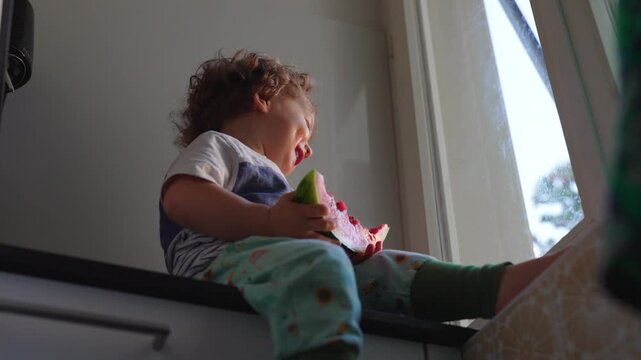 Happy toddler eating watermelon while sitting on windowsill at home, joyful childhood moment highlighting wellbeing, nutrition, playfulness and carefree development