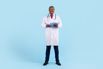 Confident mature African American doctor in white coat with stethoscope, holding clipboard and writing notes, full body on blue background © Anastasiya