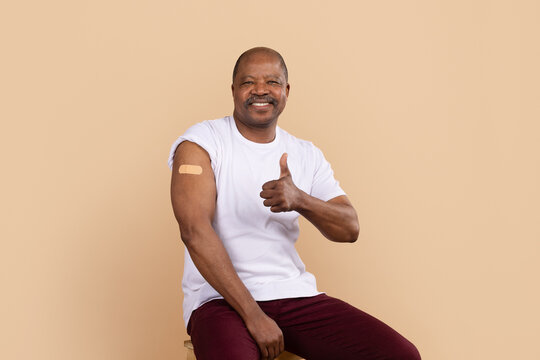 Mature African American man sitting on chair and showing vaccination bandage on arm with thumbs up gesture, smiling male posing on beige studio background