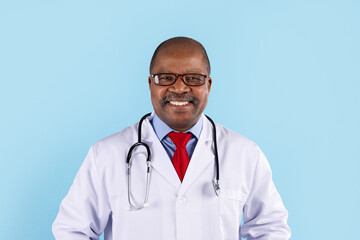 Fototapeta premium Closeup portrait of smiling mature African American doctor wearing glasses, white coat, stethoscope and red tie, blue studio background with copy space