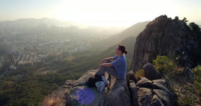 Tourist man sits on rocks at summit of Lion Rock, gazing across sweeping urban landscape of Hong Kong. Bright evening sun glows behind soft light due to some haze in air.
