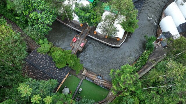 Aerial view of a glamping resort featuring white tent structures, wooden footbridges over a flowing river, lush green trees, and manicured lawns in Pangalengan, West Java, Indonesia.