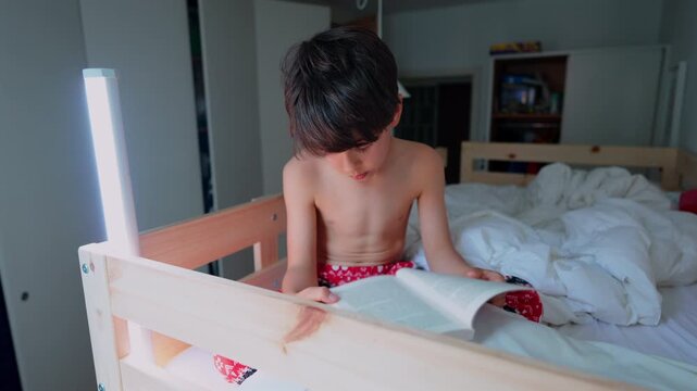 Young boy reading book while sitting on bed at home, focused childhood learning moment highlighting literacy, education, concentration and independent reading development