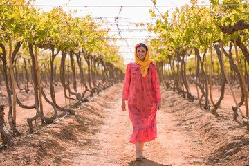 Fototapeta premium Portrait of confident Middle Eastern woman agricultural worker in traditional embroidered pink dress and yellow headscarf standing in symmetrical rows
