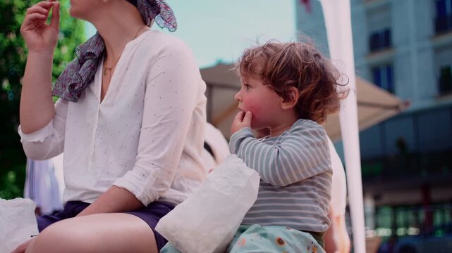 Toddler sitting beside mother outdoors while eating snack, candid family moment highlighting childhood independence, attachment, comfort and everyday connection