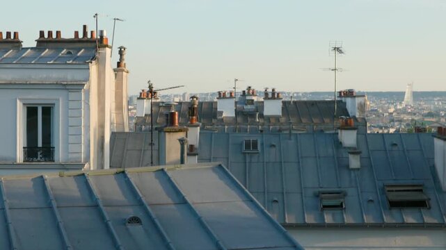 Camera tilting up over the classic zinc rooftops, antennas, and clay chimneys of Parisian apartment buildings under a clear blue sky.