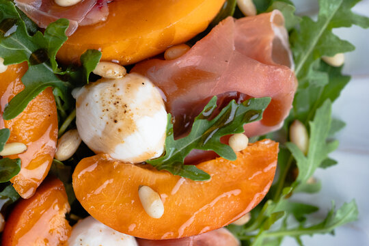 Macro close-up of a fresh summer salad featuring juicy apricot slices, creamy mozzarella pearls, delicate prosciutto, peppery arugula, and toasted pine nuts.