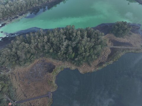 Aerial view of volcanic Dieng Plateau featuring the vibrant green waters of Telaga Warna lake surrounded by dense forest and a wooden boardwalk in Kejajar, Central Java, Indonesia.