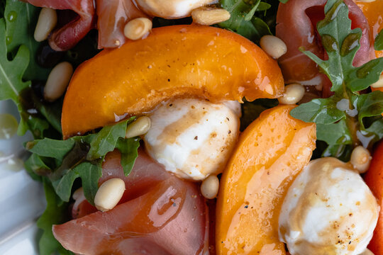 Macro close-up of a fresh summer salad featuring juicy apricot slices, creamy mozzarella pearls, delicate prosciutto, peppery arugula, and toasted pine nuts.