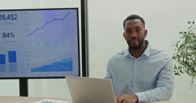 Portrait of African American bearded data analyst works on laptop in office. Financial manager predicts potential profit of deals as sits by screen with charts