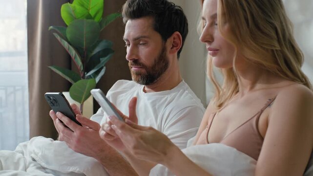 Busy family gadget addicted Caucasian couple using typing mobile phones in bed at morning woman ignoring man scrolling social media spouses concentrated browsing internet overuse smartphones problem