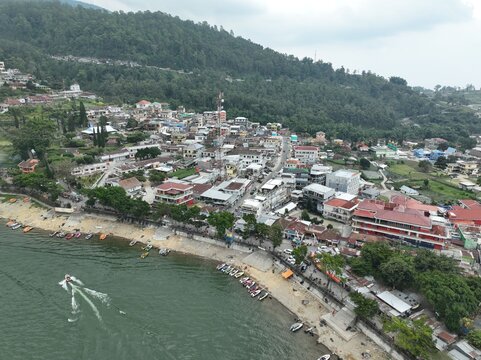Aerial view of Sarangan Lake with boats docked along the sandy shore, a motorboat on the water, and the town of Plaosan nestled against lush green hills in Plaosan, East Java, Indonesia.