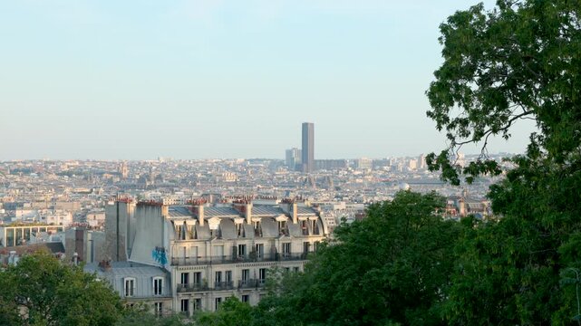 Wide panoramic view looking over the rooftops of Paris, featuring traditional mansard apartment buildings framed by leafy green trees and the Montparnasse Tower on the horizon.