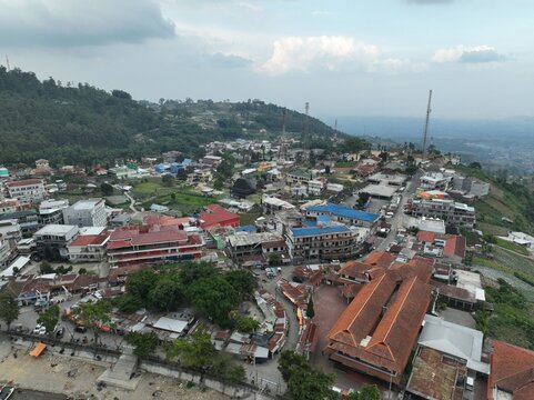 Aerial view of the town near Sarangan Lake with dense buildings, red-tiled roofs, and lush green hills under a cloudy sky in Plaosan, East Java, Indonesia.