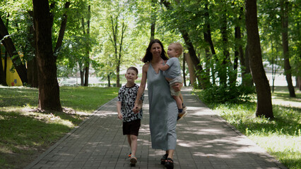Obraz premium Woman in a sleeveless dress walks down a paved park pathway holding a toddler while an older child walks beside her