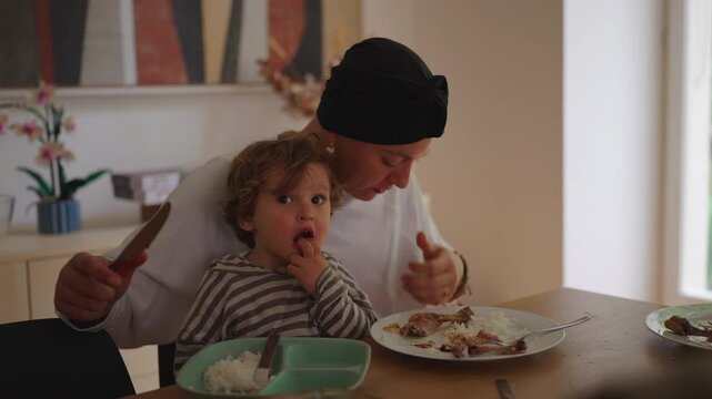 Mother eating meal while holding toddler on lap at dining table in family home during cancer recovery, highlighting caregiving, resilience, family connection and nurturing support