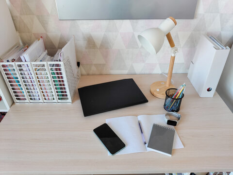 Desk with student supplies and laptop for remote learning. Workspace setup with notebook, stationery, phone, and lamp. Concept of organized study area in home interior for education.