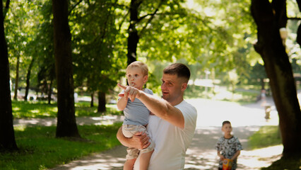 Fototapeta premium Young father holding toddler boy at shoulder height while pointing ahead along sunlit tree-lined park path; dappled afternoon light filters through leafy