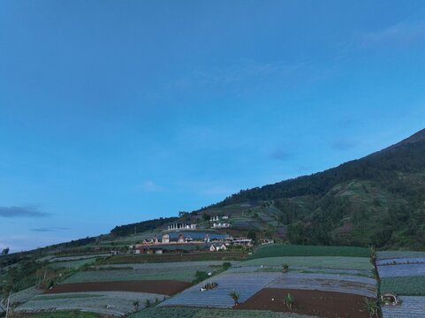 Aerial view of Linggar Jati settlement surrounded by terraced agricultural fields on the mountain slopes under a clear blue sky in Kecamatan Kaliangkrik, Jawa Tengah, Indonesia.