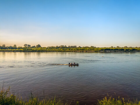 A scenic view of a motorboat with passengers traveling down the wide Vistula River, framed by wild grass on the bank in the foreground.