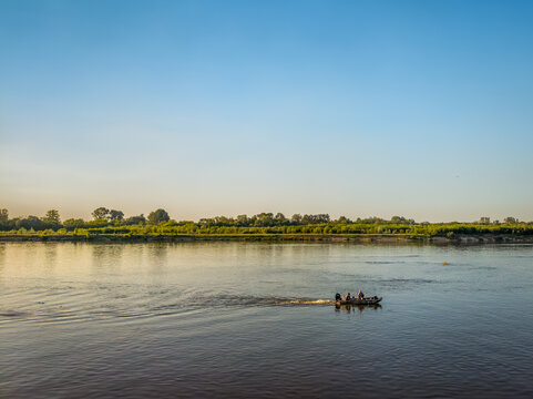 A wide landscape view of a small fishing boat cruising along the peaceful Vistula River in Poland during a bright summer afternoon.