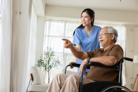 Caregiver senior wheelchair patient nurse home window pointing Asian caregiver helping elderly man in wheelchair near bright window during home health care visit